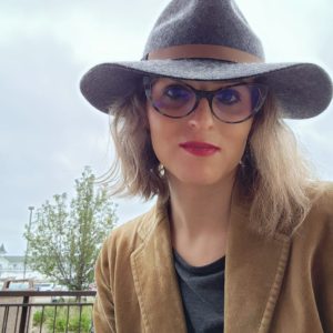 Brittyn, a white trans femme non-binary person, wears a grey wool hat with a brim on an overcast day. Brittyn also wears blue / black cats' eye glasses, has light brown hair, and keeps warm with a tannish jacket over a grey shirt. A tree is in the background.