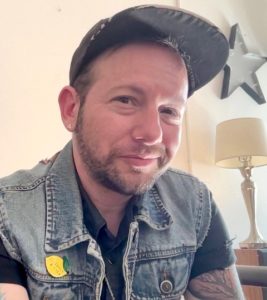Calvin grins while looking at the camera in this indoor photo. He wears a denim vest over a black shirt along with a pin and cap. In the right corner is a lamp and a star on the wall.